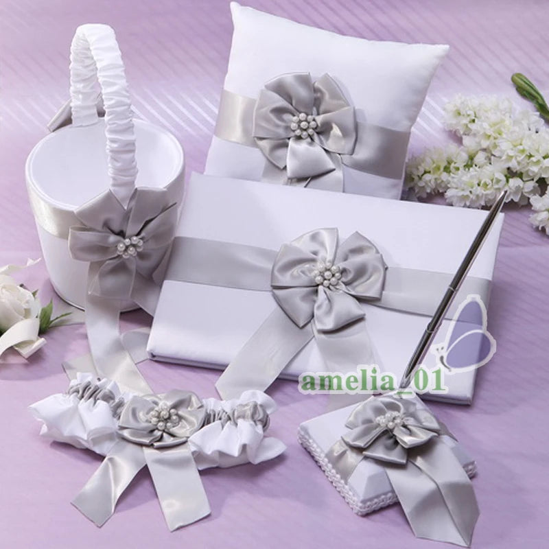 Elegant wedding accessory set on a purple surface includes a pillow, box, pen, and basket, all adorned with silver-gray satin bows and pearly accents, surrounded by white flowers.