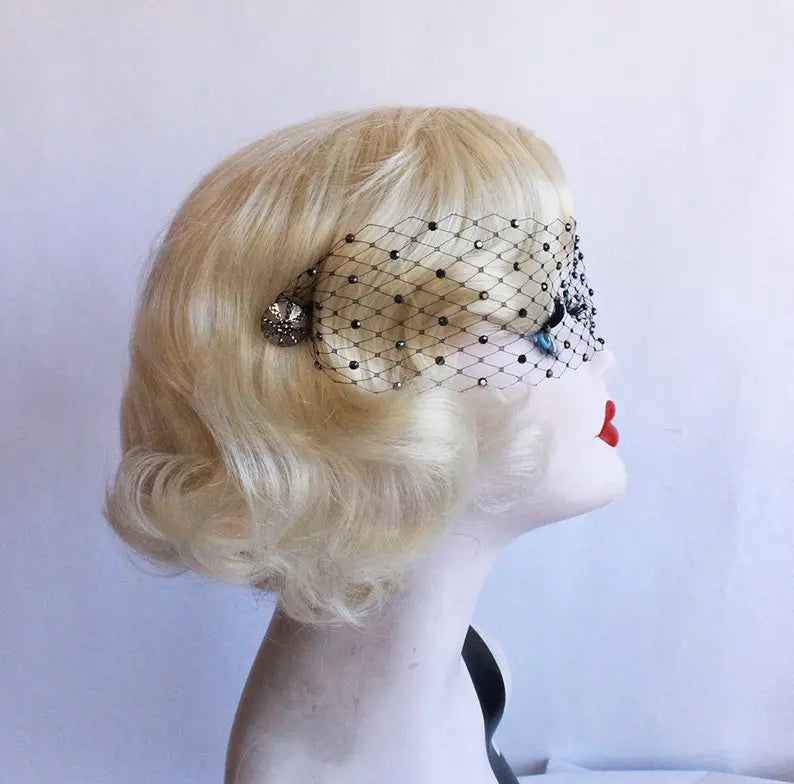 Blonde mannequin head with retro waves and red lips, wearing a black netted veil adorned with a jeweled brooch, set against a plain background.
