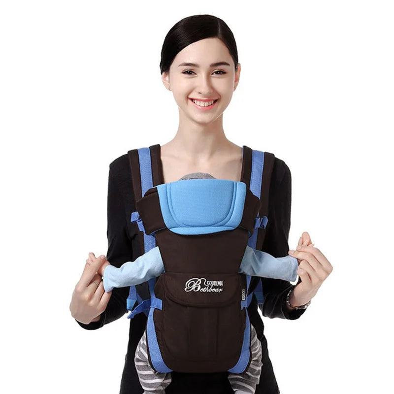 A woman smiling while wearing a black and blue baby carrier, holding a baby dressed in blue and gray. The baby is nestled securely with a padded headrest.