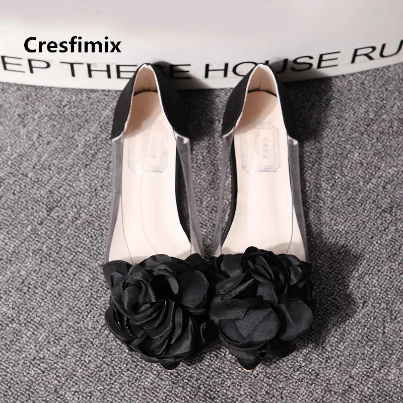 Elegant black open-toe shoes with large floral embellishments on the front, displayed on a textured gray carpet.
