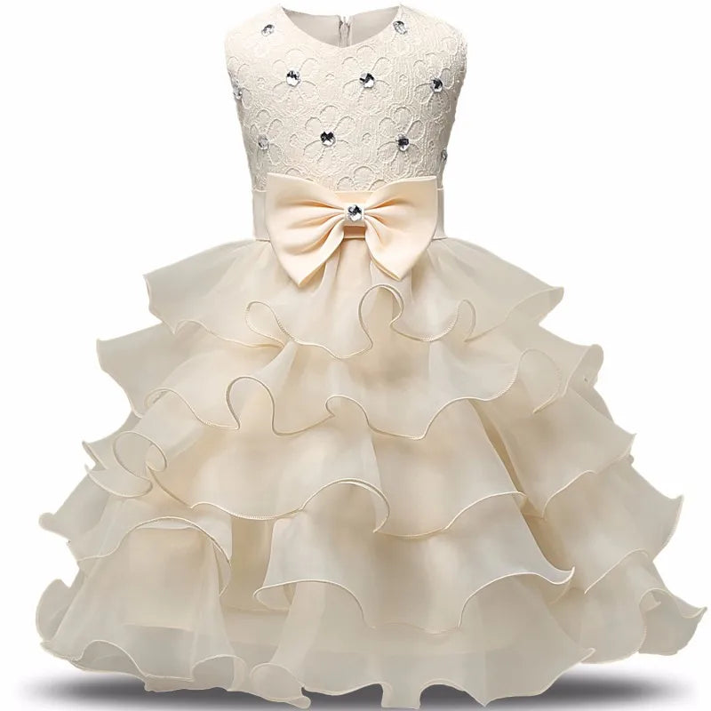 Elegant cream-colored dress for a child, featuring a lace bodice adorned with small gems, a large bow at the waist, and a voluminous tiered ruffle skirt.