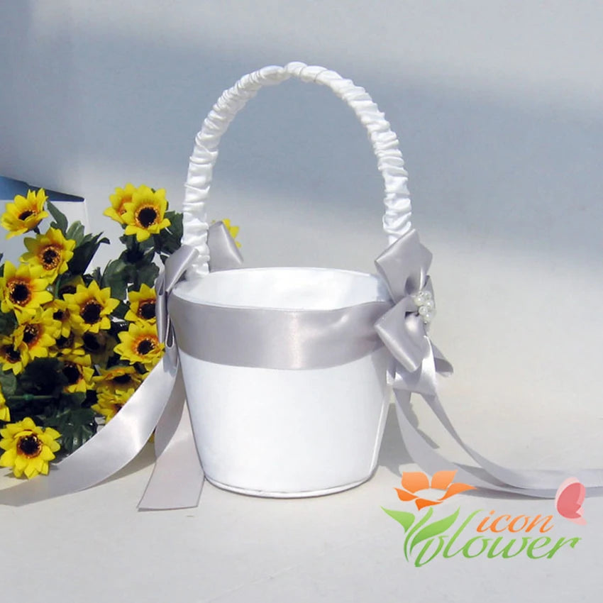 White satin flower girl basket with silver ribbon and bow, embellished with pearls, placed beside a bouquet of bright yellow flowers.