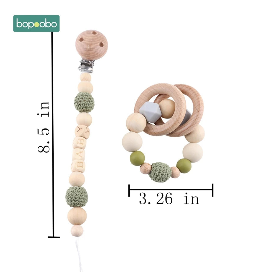 Wooden pacifier clip and teether set with green crochet beads, beige and olive accents. Clip measures 8.5 inches, teether is 3.26 inches. Brand logo in top left.