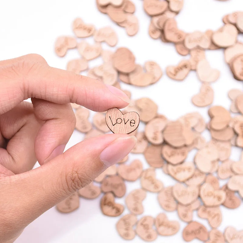 A hand holds a small wooden heart engraved with "Love," with more similar hearts scattered in the blurred background.