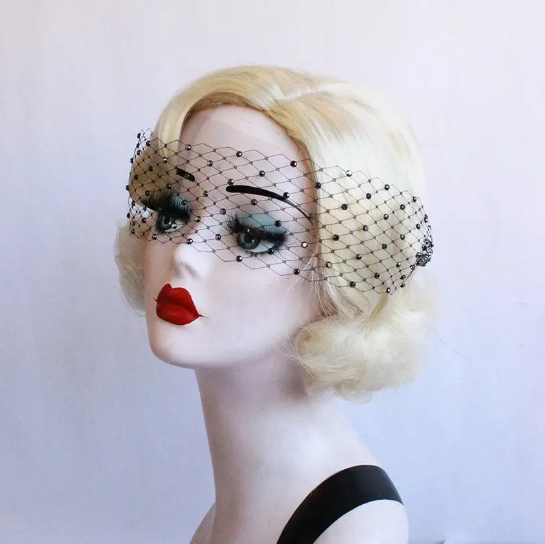 A mannequin head with blonde hair wears a black netted veil adorned with small beads. The mannequin has bold red lips and dramatic eye makeup. The background is plain white.