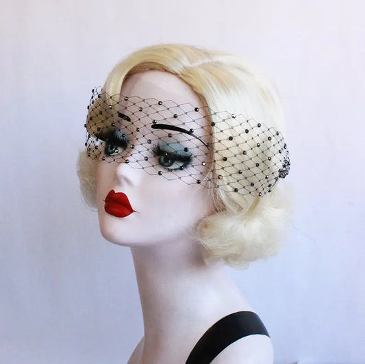 A mannequin head with blonde hair wears a black netted veil adorned with small beads. The mannequin has bold red lips and dramatic eye makeup. The background is plain white.