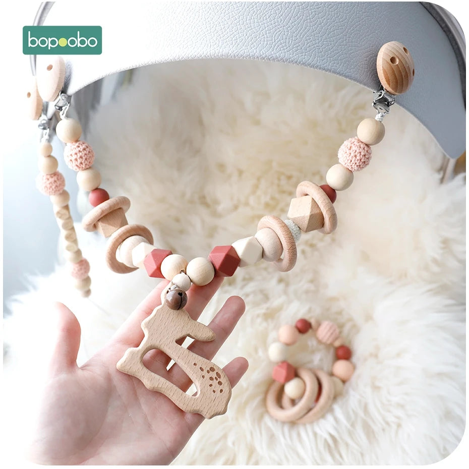 Wooden baby toy with beads, rings, and a dinosaur shape, mounted on a stroller. A hand holds part of the toy. Background is fluffy white fabric. Small matching bracelet nearby.