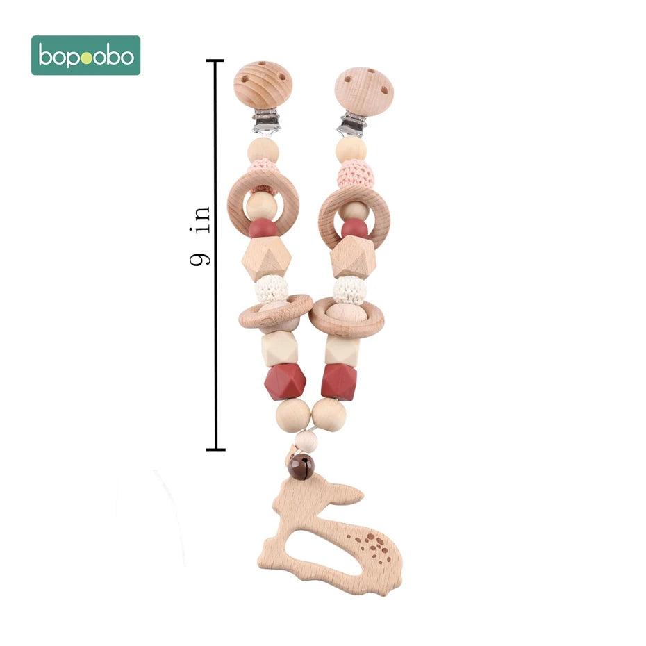 Wooden pacifier clip with beads, rings, and a bunny-shaped teether. Dual clip design, 9 inches long. Multicolored with natural and red hues. Brand "bopoobo" in corner.