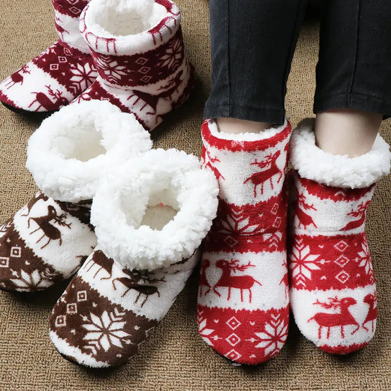 Fluffy indoor slippers with festive red and brown patterns featuring reindeer and snowflakes, placed on a carpet. A person wears the red pair, showcasing cozy white lining.