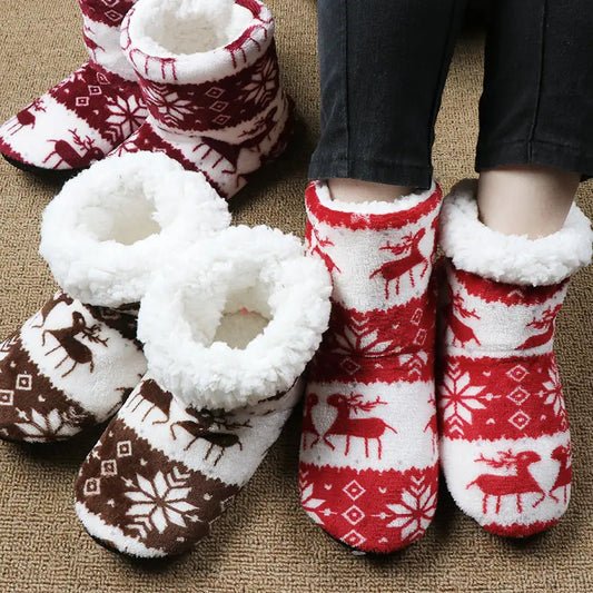 Fluffy indoor slippers with festive red and brown patterns featuring reindeer and snowflakes, placed on a carpet. A person wears the red pair, showcasing cozy white lining.