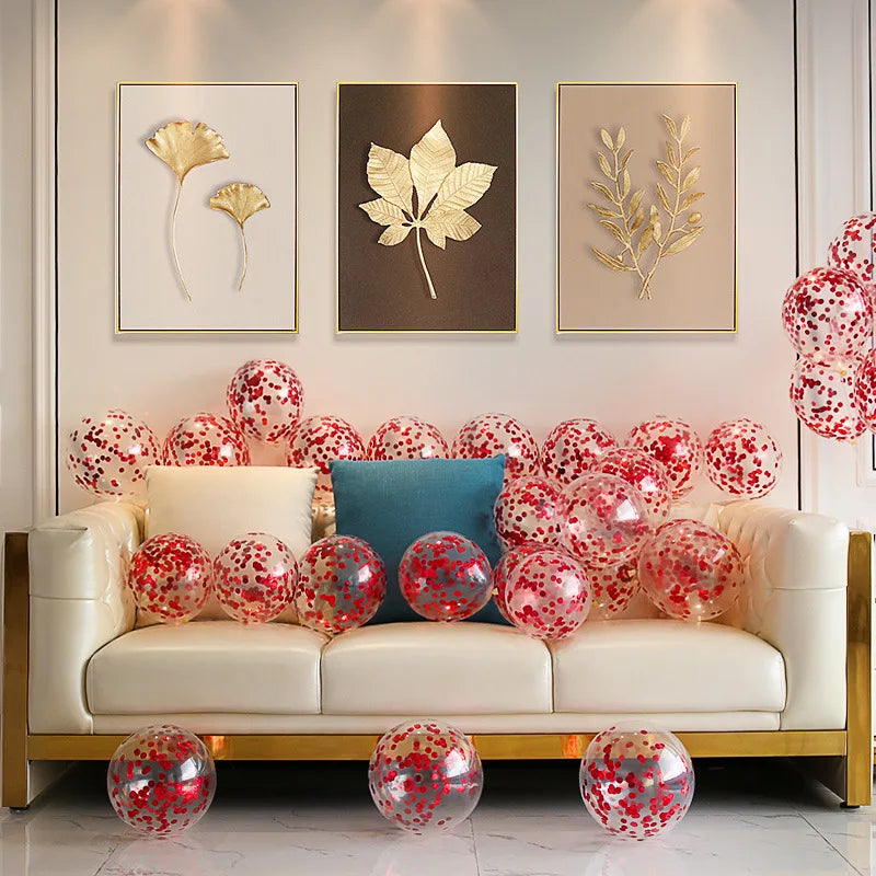 Sleek white couch with a blue cushion, surrounded by transparent balloons with red confetti. Above, three framed gold leaf artworks hang on a softly lit wall.