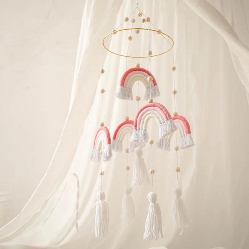 A decorative mobile with hanging pink and white woven rainbows and tassels, accented by wooden beads, suspended against a soft, cream-colored fabric backdrop.