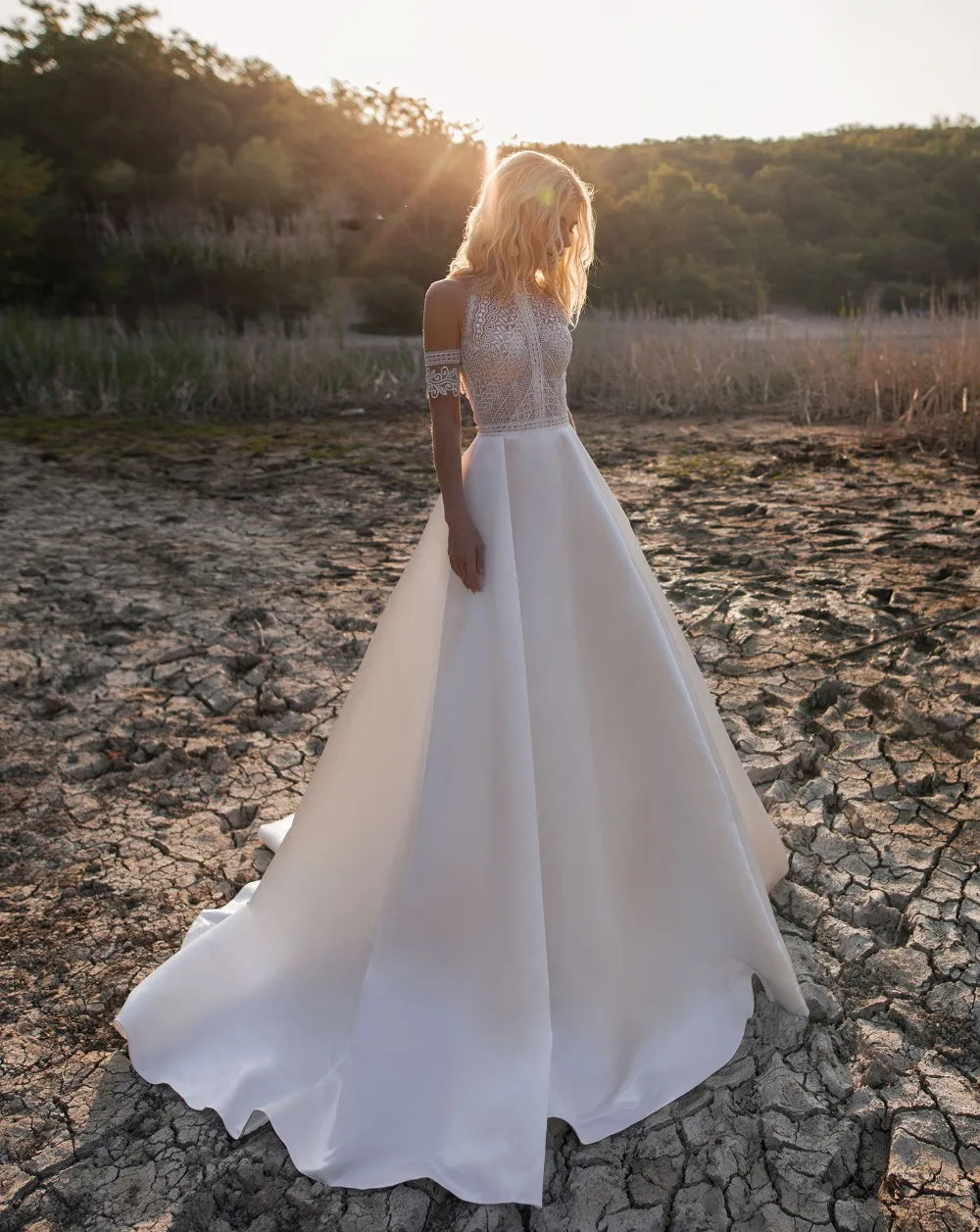 A woman in a lace wedding dress with a flowing skirt stands on cracked earth, bathed in sunset light, with trees in the background.