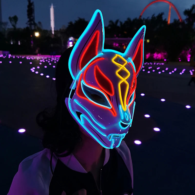 A person wearing an LED fox mask with vibrant blue, red, and yellow lights stands in a dimly lit outdoor area with glowing purple lights scattered on the ground.