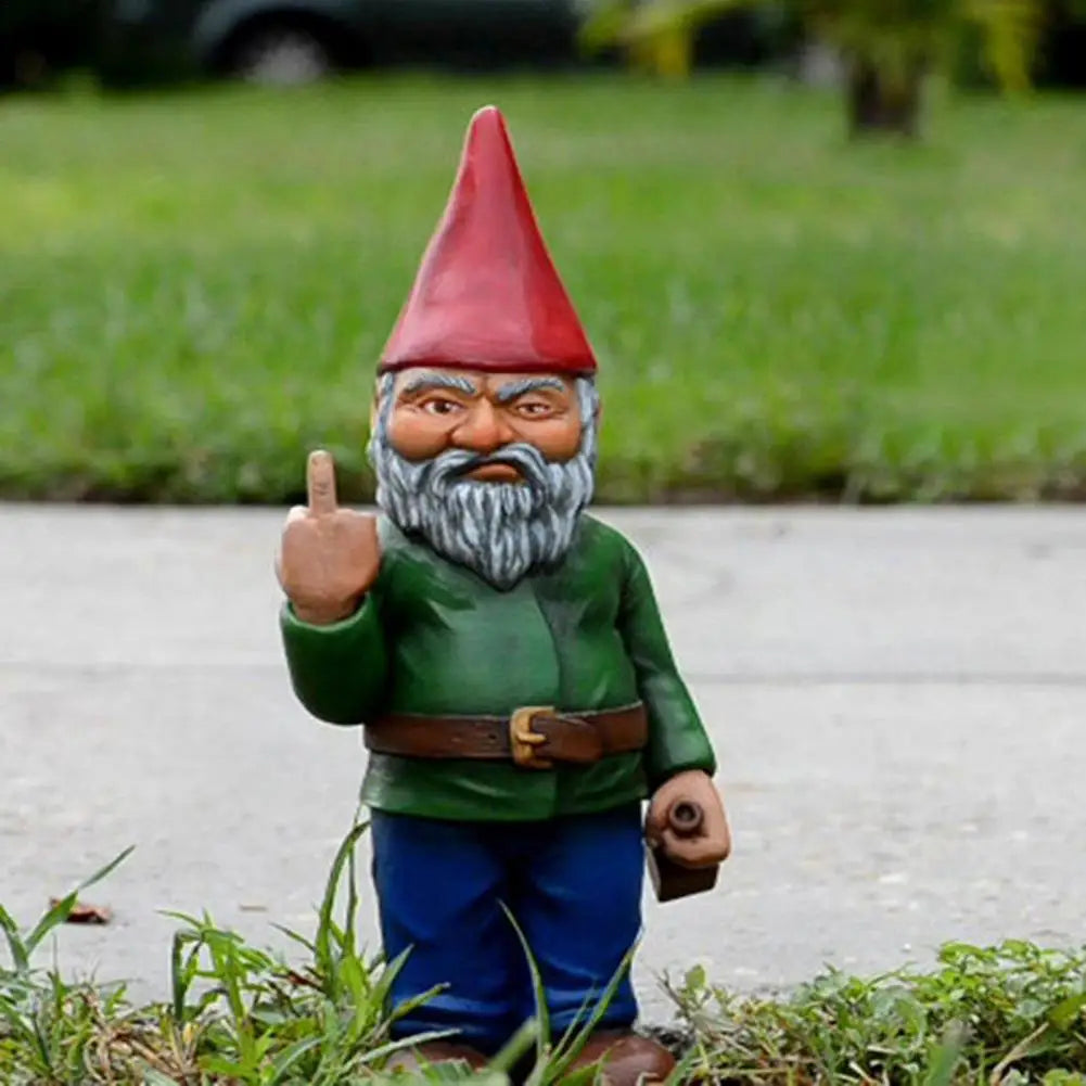 Garden gnome with a red hat and a green shirt, holding a middle finger up. It stands on grass with a stern expression, next to a sidewalk.