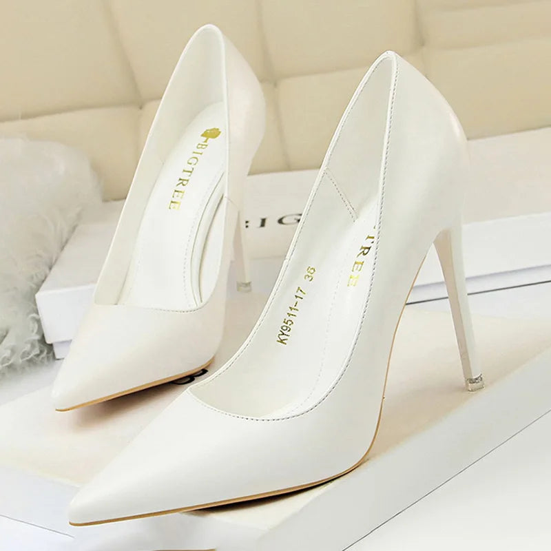 Elegant white stiletto heels with pointed toes, displayed on a beige surface. The shoes have branding inside and are designed with slender, high heels.