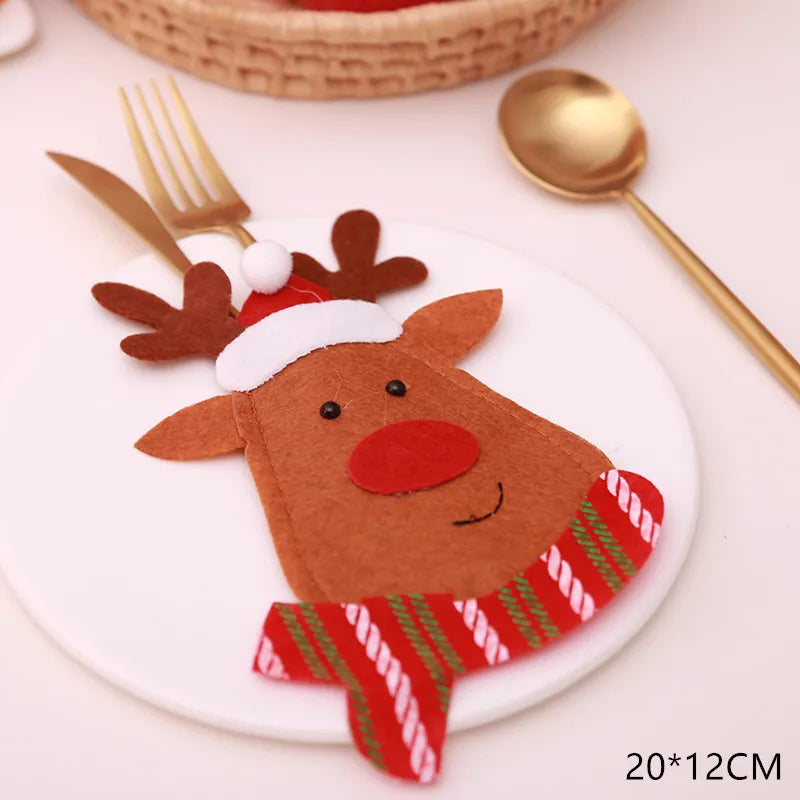 Festive table setting with a reindeer-shaped felt cutlery holder on a white plate, adorned with a Santa hat and red scarf. Gold fork and spoon beside it. Dimensions: 20x12cm.