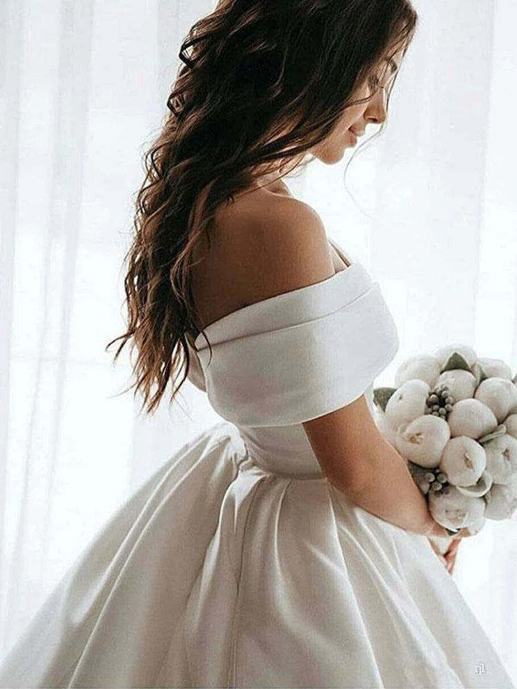 Bride in an off-the-shoulder white dress holding a bouquet of white flowers, standing against sheer curtains with long wavy hair cascading down her back.