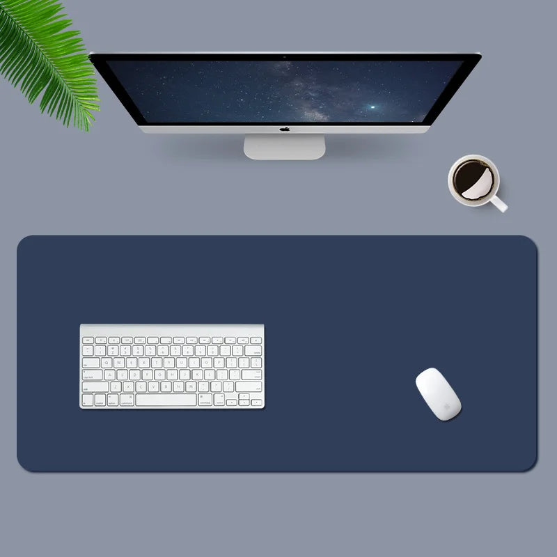 A clean and minimalist desk setup with a large monitor displaying a starry sky, a keyboard, mouse on a dark mat, a cup of coffee, and a green leaf on a grey background.