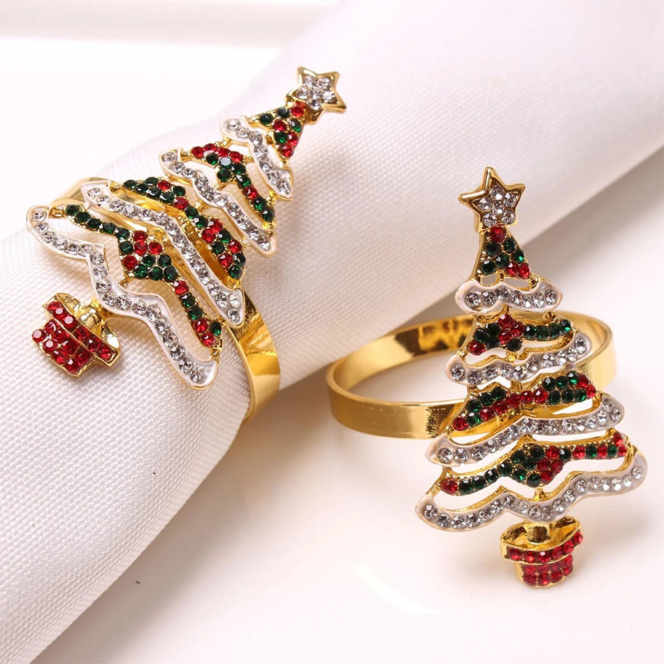 Gold napkin rings shaped like Christmas trees, decorated with red, green, and white rhinestones and topped with a star, elegantly displayed on a rolled white napkin.