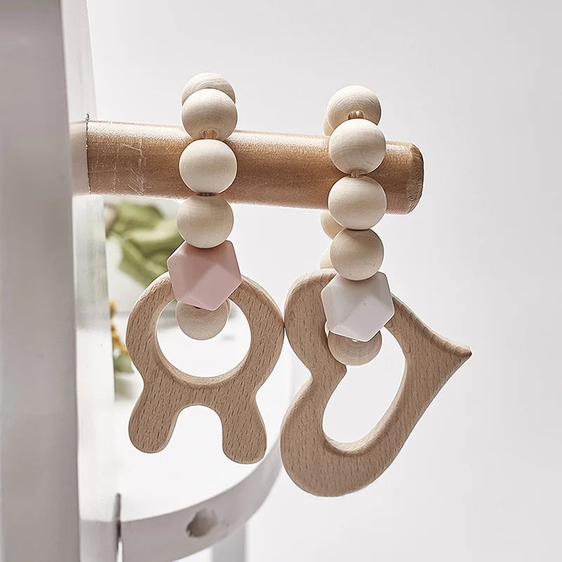 Two wooden teething rings hang from a rail, featuring light wooden beads and hexagonal silicone beads in pink and white. The rings have playful, abstract shapes.