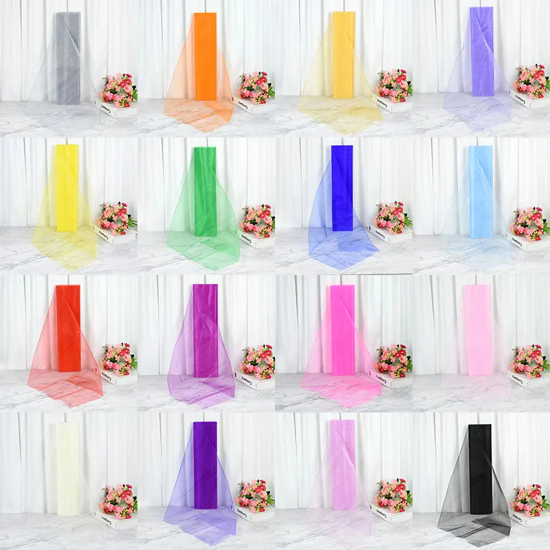 Colorful rolls of tulle fabric in 16 shades, from gray to black, displayed against a white curtain backdrop with flower arrangements beside each roll.