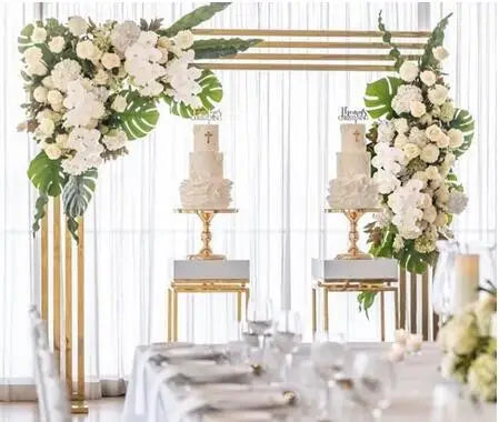 Elegant event setup with two tiered cakes on gold stands under a floral arch of white roses and greenery. White draped background and table set with crystal glassware in the foreground.