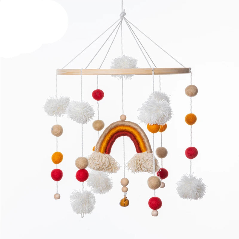 Colorful handmade baby mobile with pom-poms and a rainbow motif, hanging from a wooden ring by strands. Features red, orange, beige, and white elements on a white background.