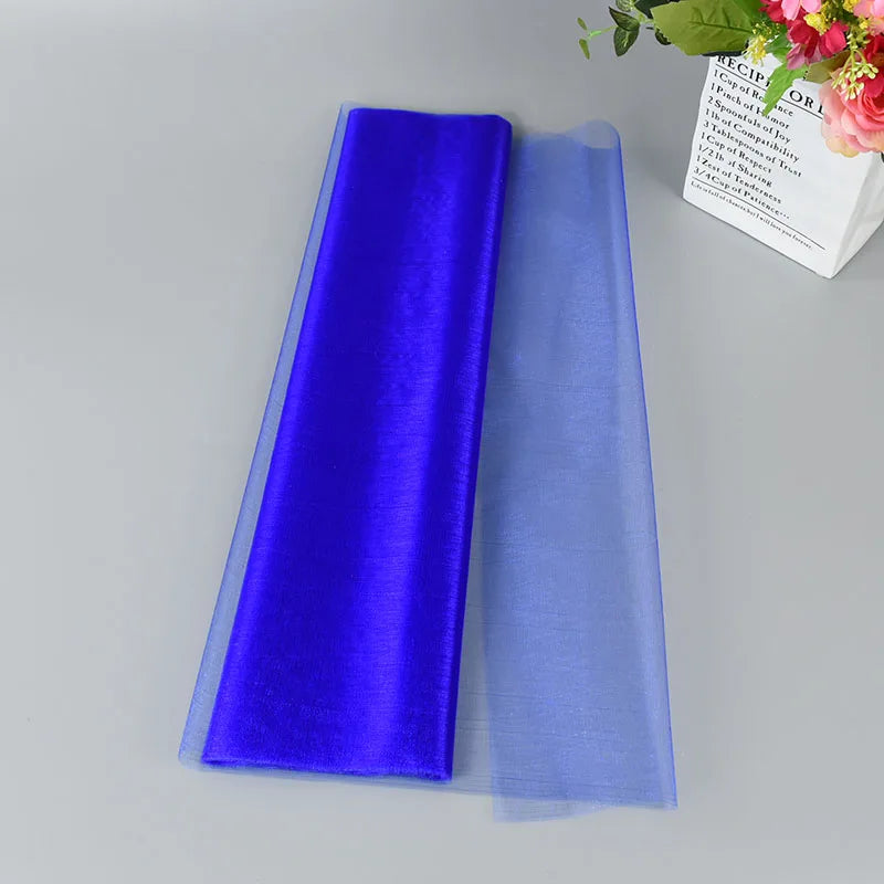 Rich blue and translucent purple tulle fabric laid flat on a gray surface, with a bouquet of pink flowers and green leaves in a box visible in the upper right corner.