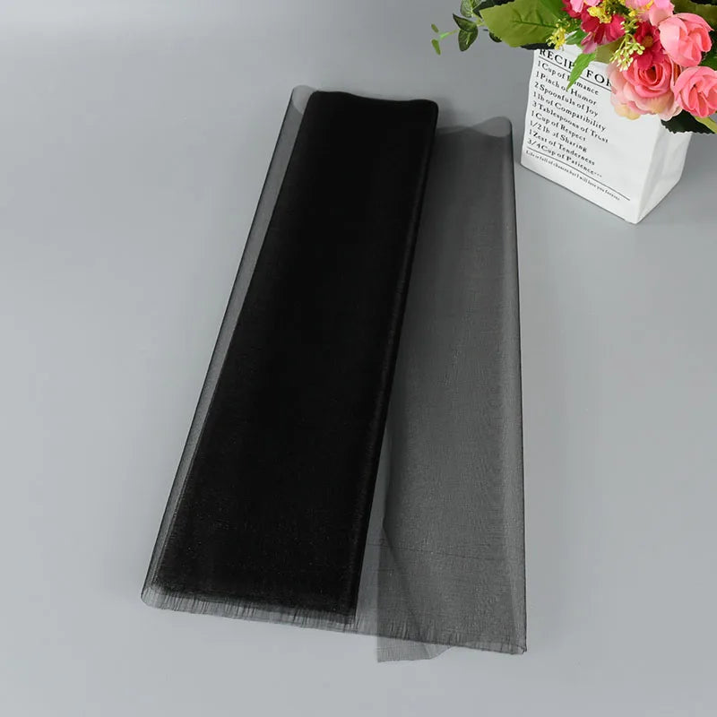 A black, sheer tulle fabric lies flat on a gray surface beside a white vase holding vibrant pink flowers.