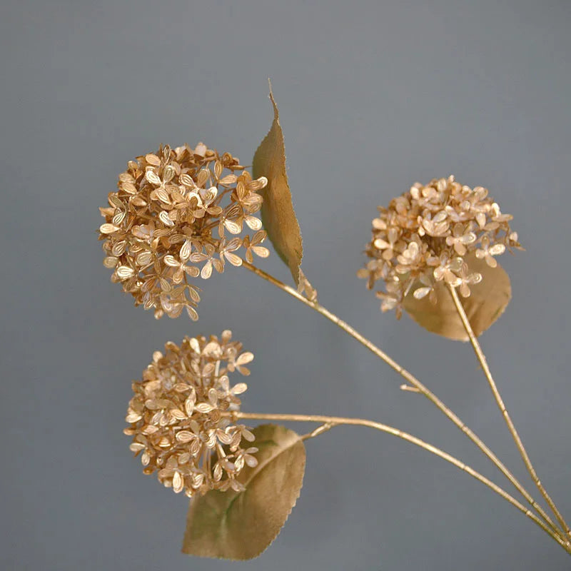 Golden, dried hydrangea flowers with delicate petals and leaves against a soft gray background, elegantly arranged on thin stems.