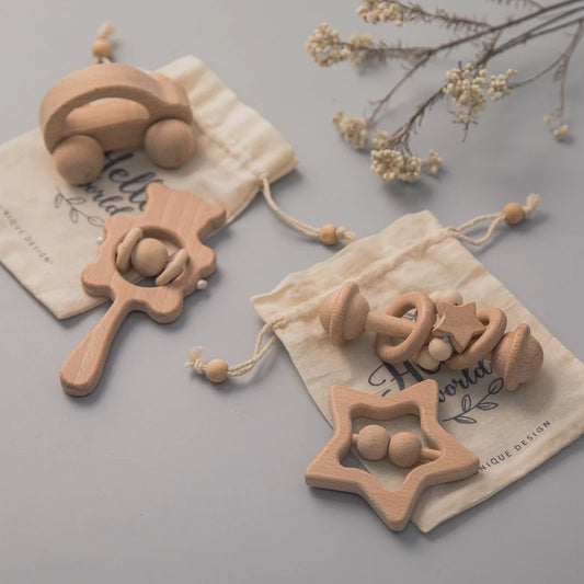Wooden baby toys on a table, including a car, a rattle, and a star-shaped teether, placed on two drawstring bags with delicate dried flowers in the background.