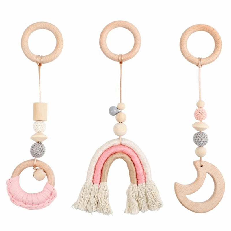 Three wooden hanging toys: a ring with pink yarn, a pink-and-cream rainbow, and a crescent moon. Each includes beads and hangs from a looped string.