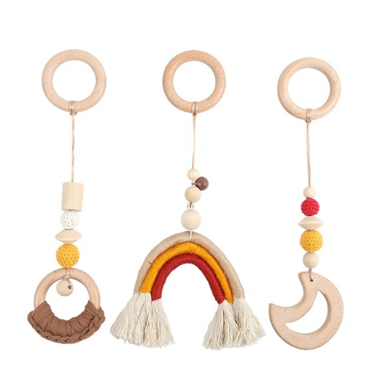 Three wooden baby teething toys with rings, featuring colorful beads and shapes: a crescent moon, rainbow, and braided loop, hanging from cords on a white background.