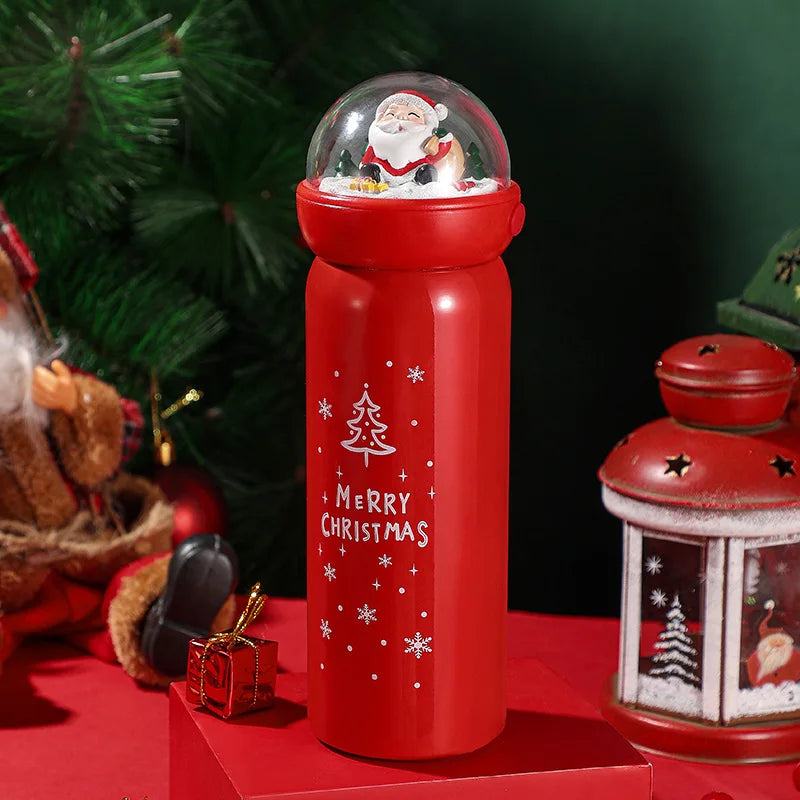 Red Christmas thermos with a Santa figurine on top, snowflakes, and the words "Merry Christmas." Surrounded by festive decor, including a small gift and a lantern.