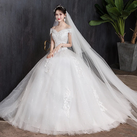 Bride in an off-the-shoulder white wedding gown with lace details and a long flowing veil, standing by a dark wall and large potted plant.