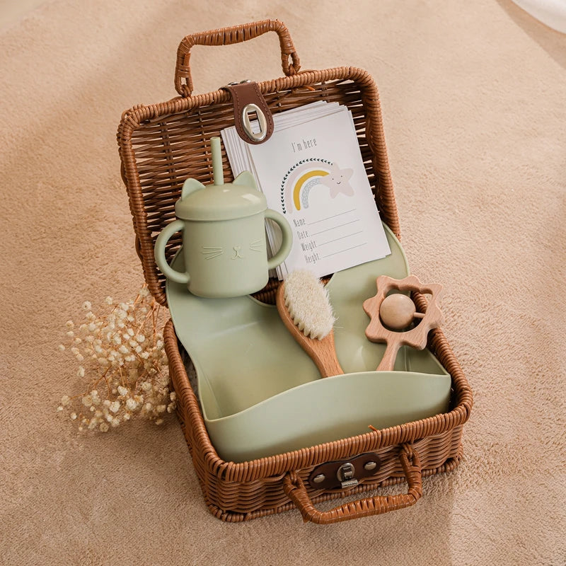 A wicker basket contains a green cat-shaped sippy cup, a baby brush, a teether, and a printed card with a rainbow design. The basket is placed on a beige textured surface.