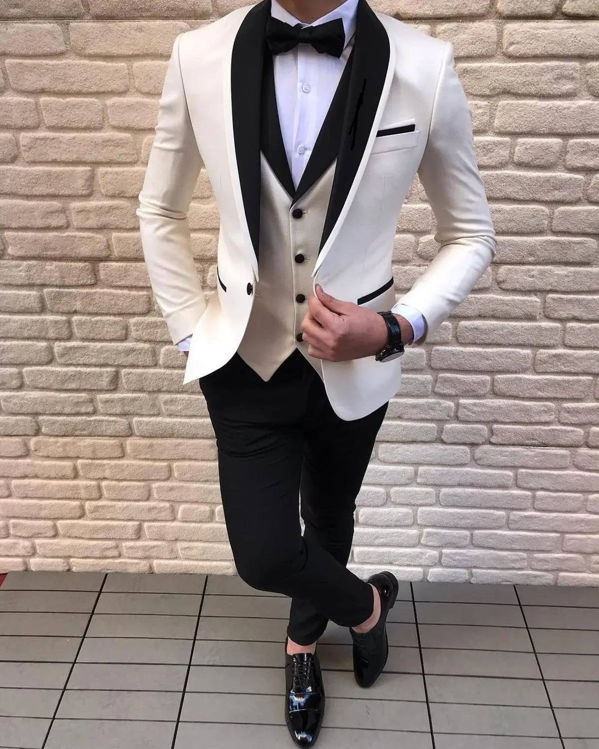 Man in a white tuxedo with a black lapel, black trousers, and shiny black dress shoes. He stands cross-legged against a light brick wall, hand adjusting the jacket.