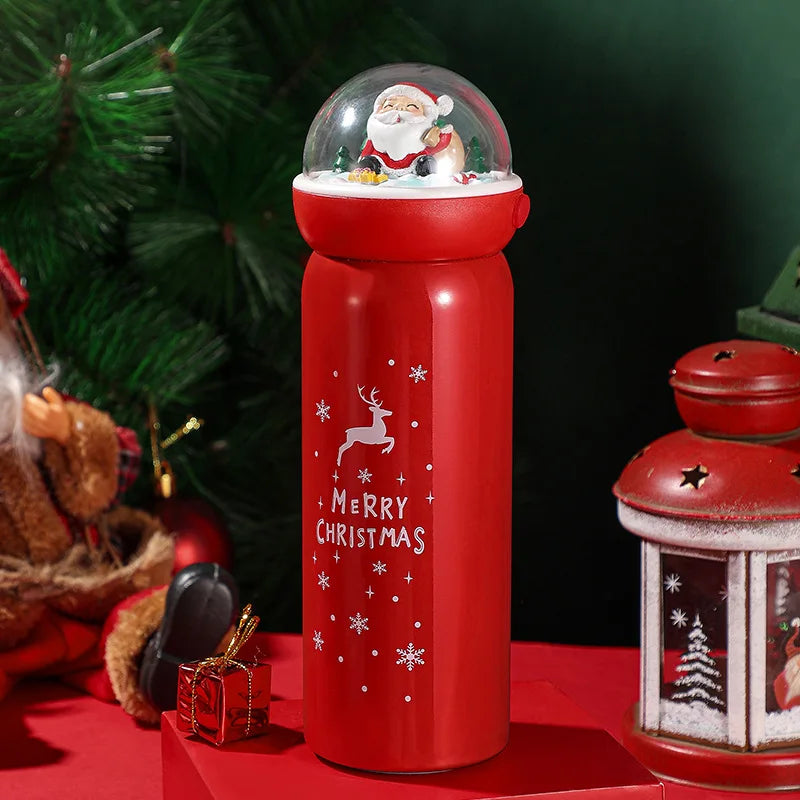 Red Christmas-themed thermos with Santa snow globe lid and "Merry Christmas" text. Decorated with snowflakes and reindeer, surrounded by festive decor and a small gift box.