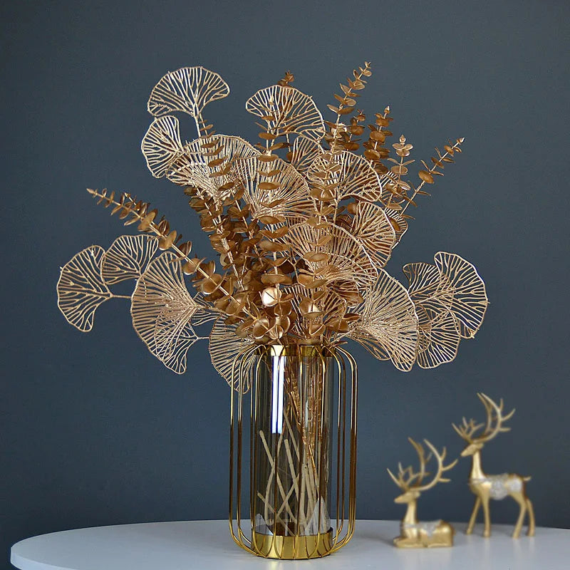 Golden decorative leaves and branches in a tall, transparent vase with a golden grid design on a white surface. Two small golden deer figurines sit beside it against a dark background.