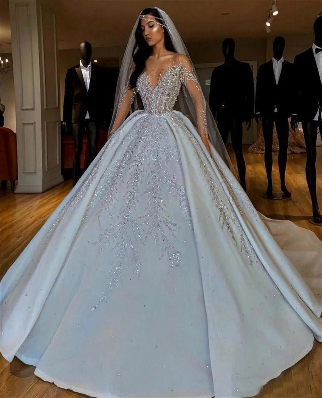 A woman in a sparkling, off-the-shoulder ball gown with a fitted bodice and long veil stands indoors. Mannequins with suits are visible in the background under soft lighting.