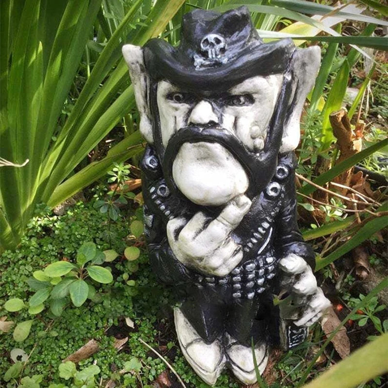 A garden gnome with a biker outfit, scowling expression, and skull-adorned hat, stands among greenery giving a rude gesture with one hand.