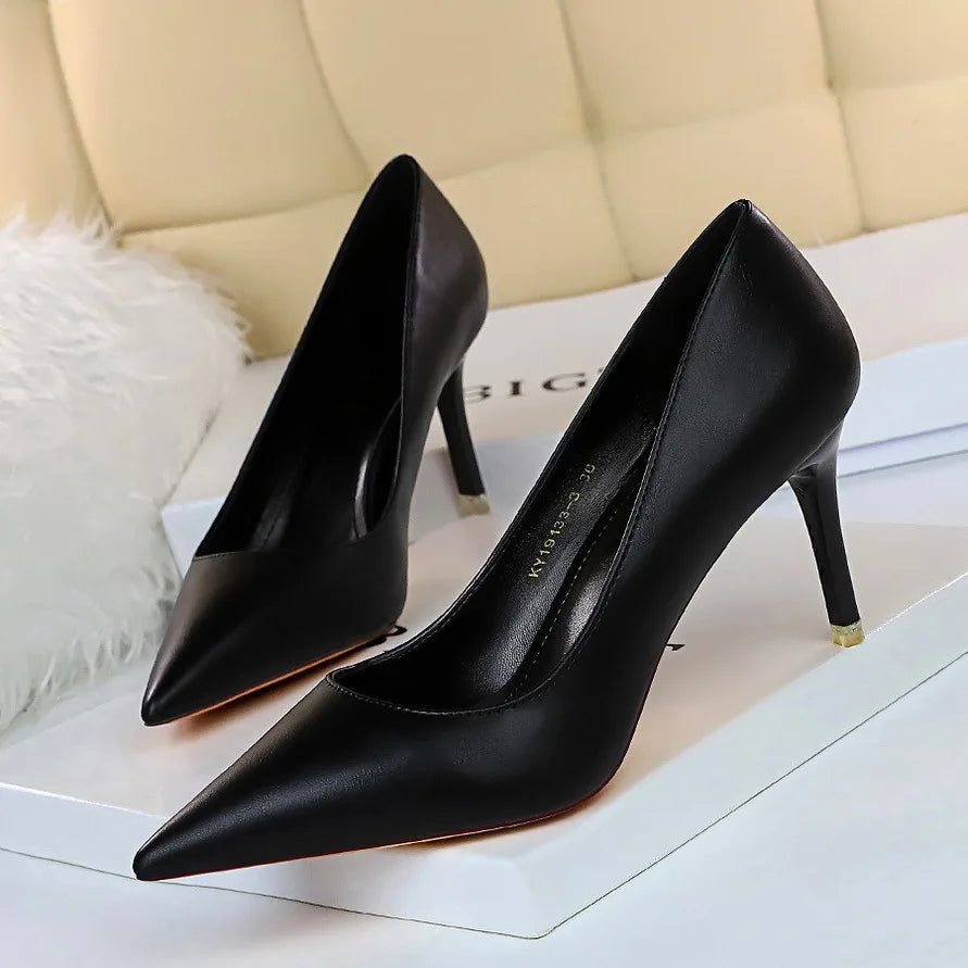 Elegant black pointed-toe stilettos with a sleek design, displayed on a white surface. Background features a white fluffy pillow and beige cushioned seat.