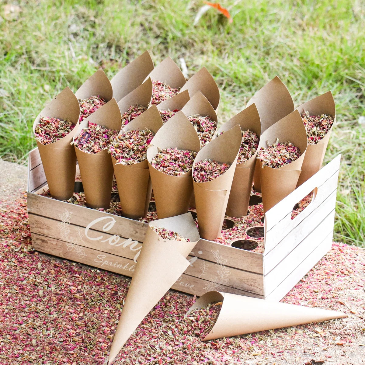 Brown paper cones filled with colorful dried flower petals are arranged in a wooden box on grass. Some petals are scattered on the ground around the box.
