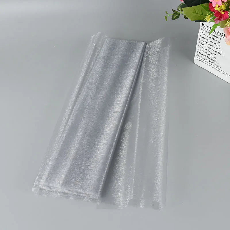 Silver sheer organza fabric neatly folded on a gray surface, with a decorative bouquet of flowers in the background.