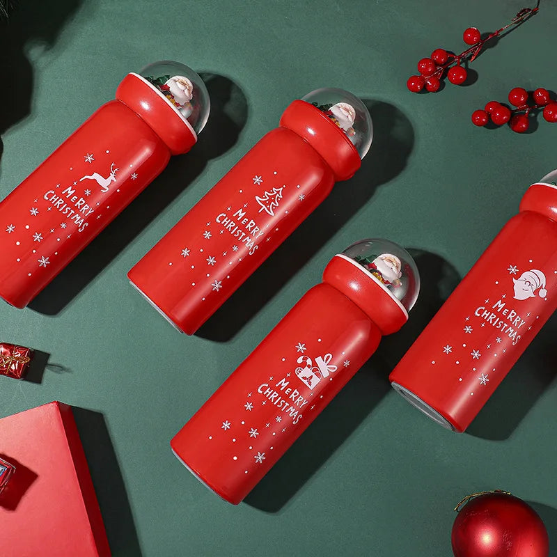 Four red bottles with "Merry Christmas" and festive designs, topped with snow globe caps featuring Santa Claus, set on a green surface with red berries and ornaments.