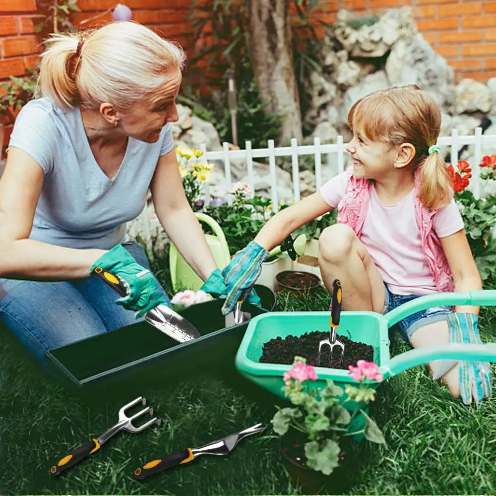 A woman and a child are gardening together, using tools and a wheelbarrow filled with soil. They are surrounded by blooming flowers, a white fence, and a brick wall in the background.