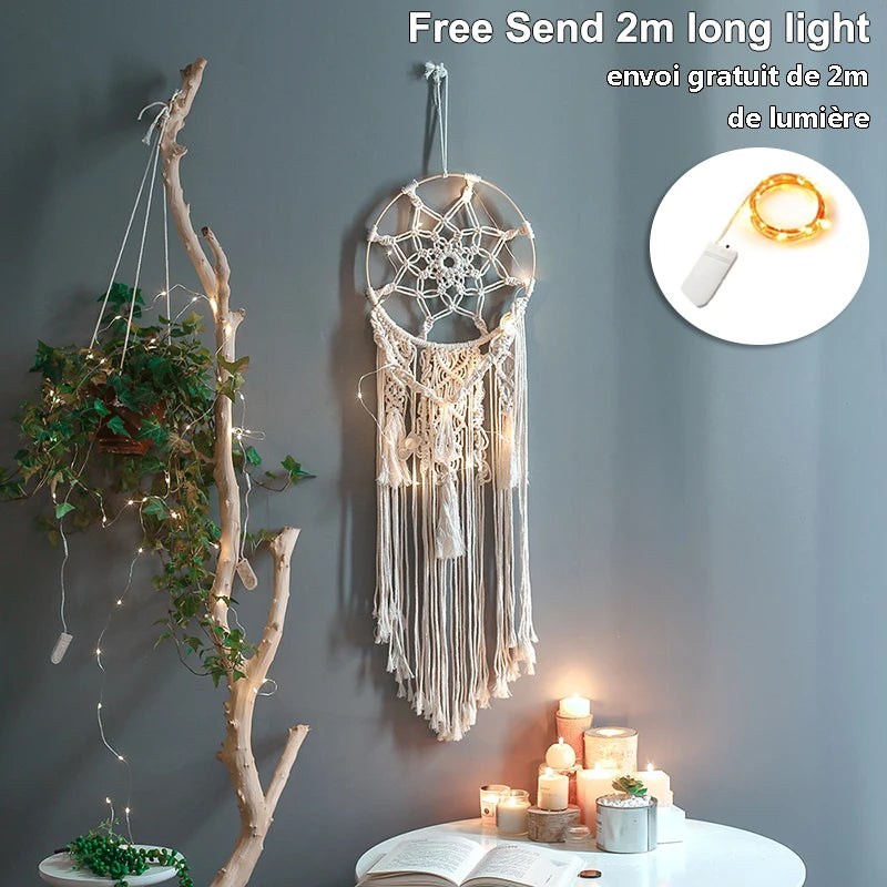 Macrame dream catcher with fairy lights hangs on a grey wall. A small shelf with a plant in strings is nearby, and a table below features lit candles and an open book.