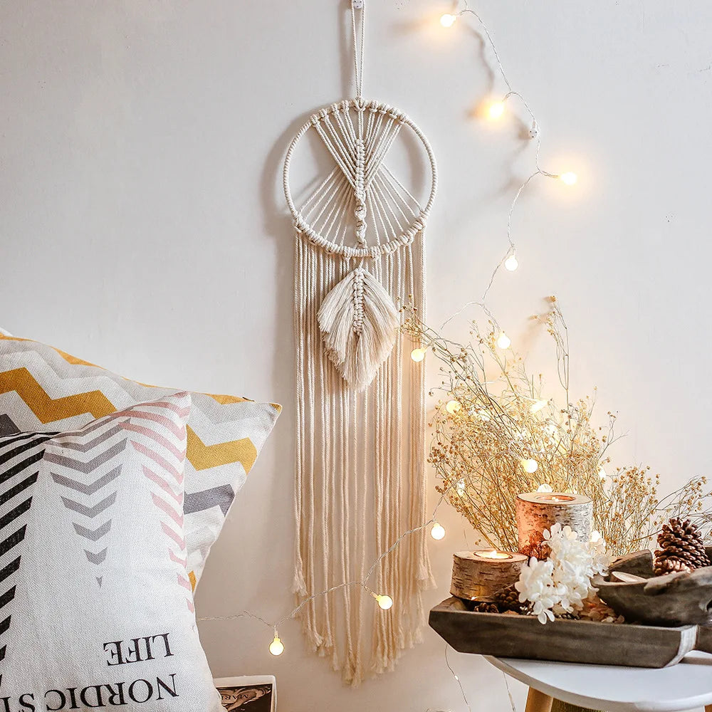 Macramé wall hanging with leaf design, framed by string lights, beside pillows with chevron patterns. A table holds birch candles, dried flowers, and pinecones. Cozy decor vibe.
