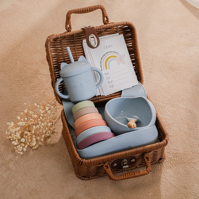 A wicker basket contains a set of pastel-toned baby dishes, a sippy cup with ears, and a baby announcement card with rainbow design on a soft beige surface.